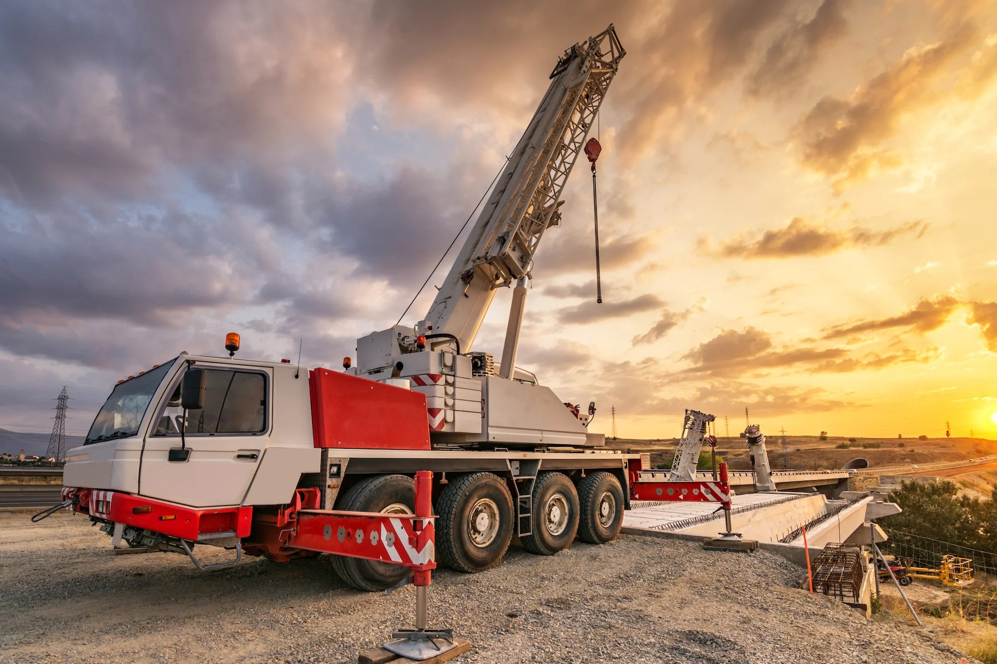 A large mobile crane with an extended boom is stationed on a construction site at sunset, with the sky partly cloudy and beams of sunlight in the background.