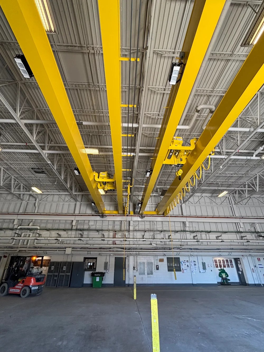 Interior of an industrial warehouse with yellow overhead cranes attached to the ceiling. The spacious area has concrete floors, machinery, and vehicles visible along the walls. Bright lighting illuminates the space.