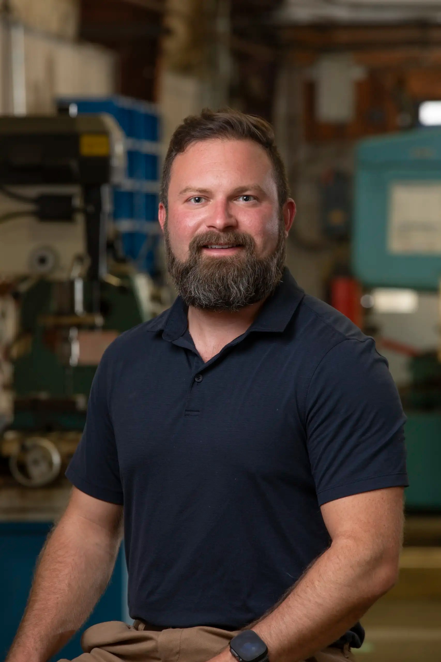 A bearded man wearing a dark polo shirt and watch poses and smiles in an industrial workshop setting, with machinery and tools visible in the background.