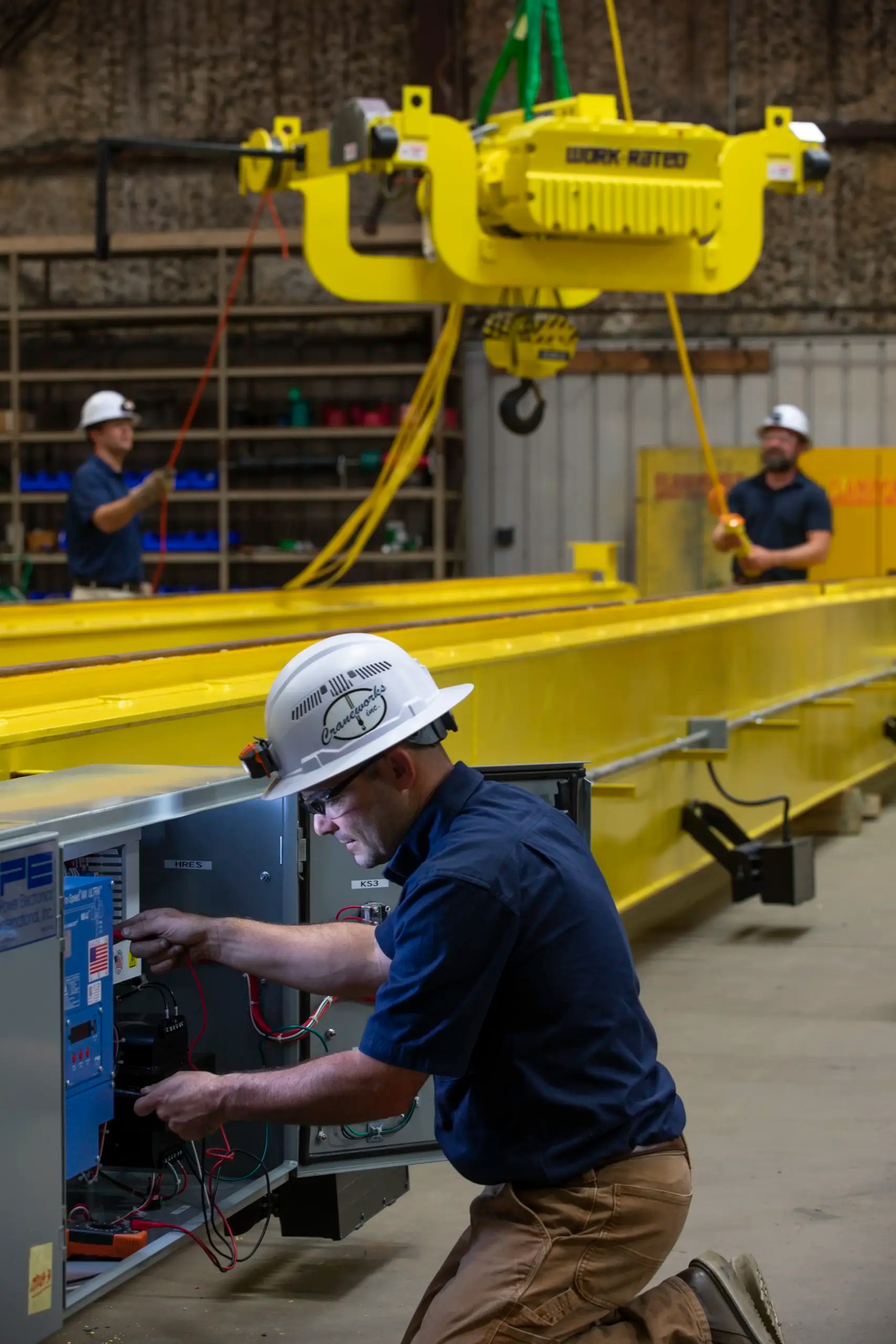 A worker in a hard hat adjusts wiring inside a control panel near a large yellow industrial crane, while two other workers operate the crane in the background inside a warehouse.