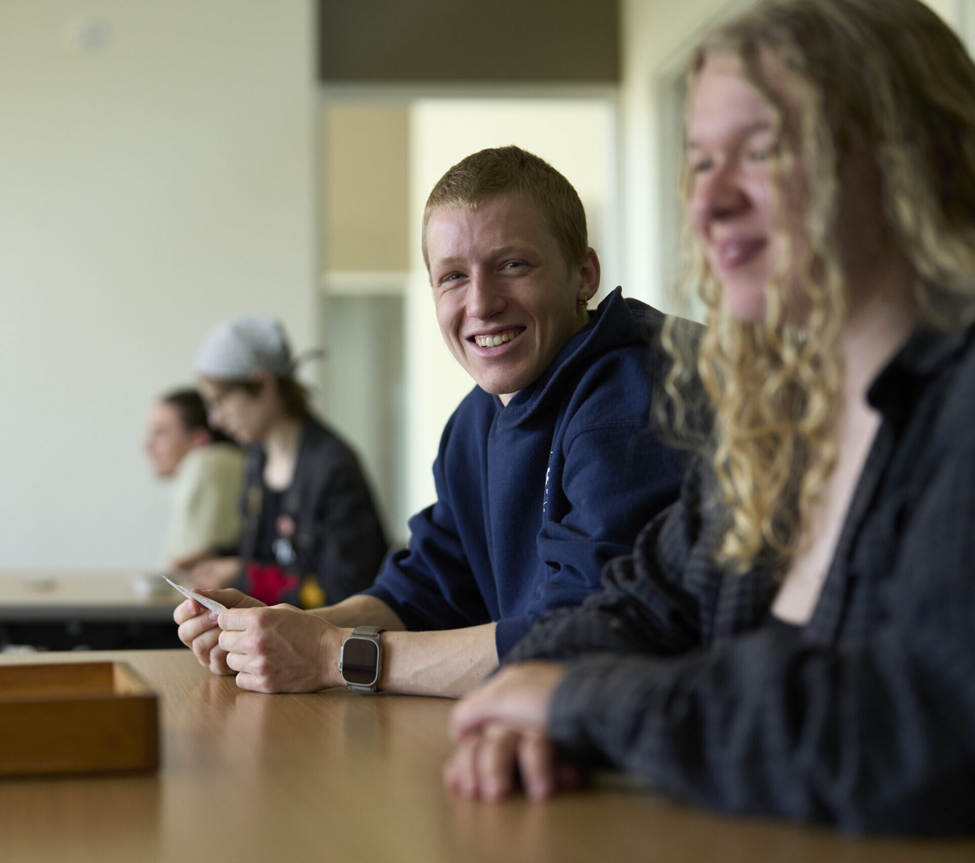 A group of students sit in a classroom. The one student in focus is smiling and holding a piece of paper.