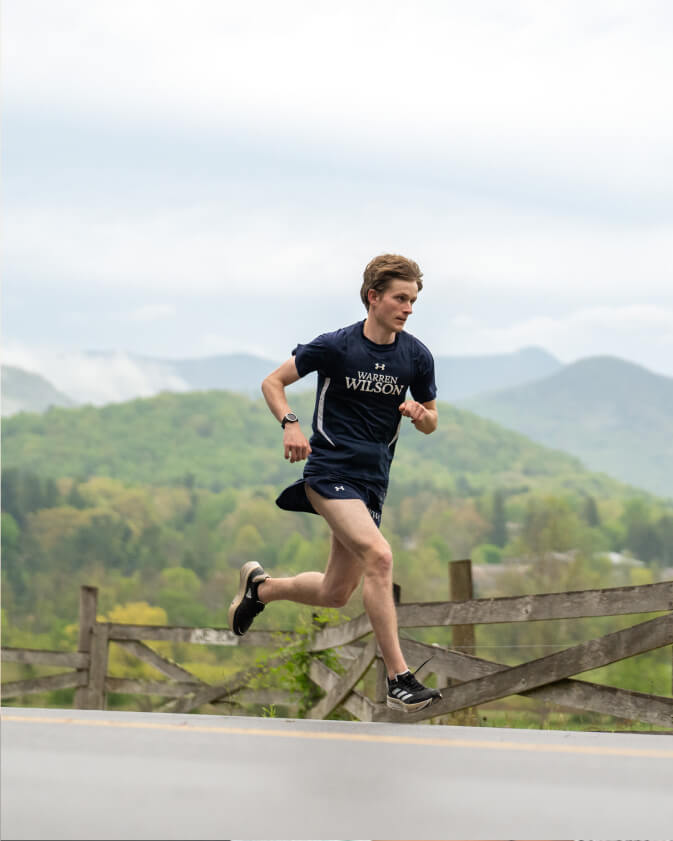 A cross-country student runs along a road. Behind them is a beautiful mountain range.
