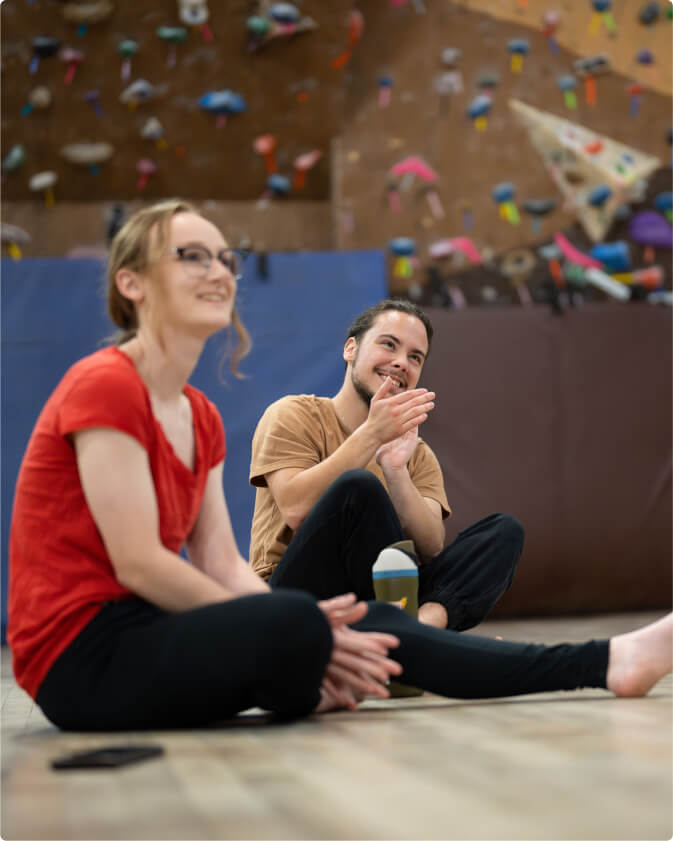 Two students sit in the floor of bryson gym watching a peer perform on aerial silks. Both are smiling and clapping.