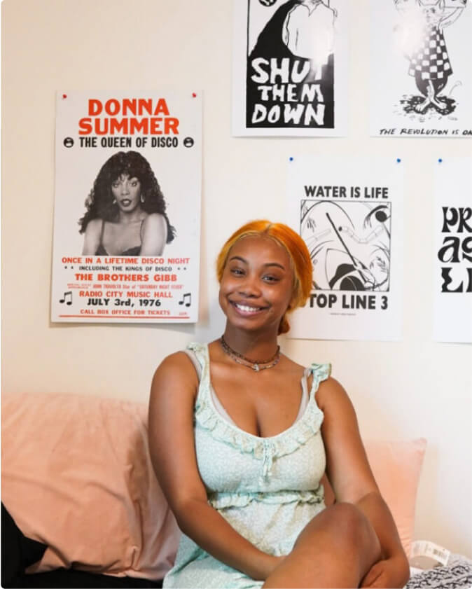 A student sits on their bed in their dorm. On the wall behind them are posters that say 