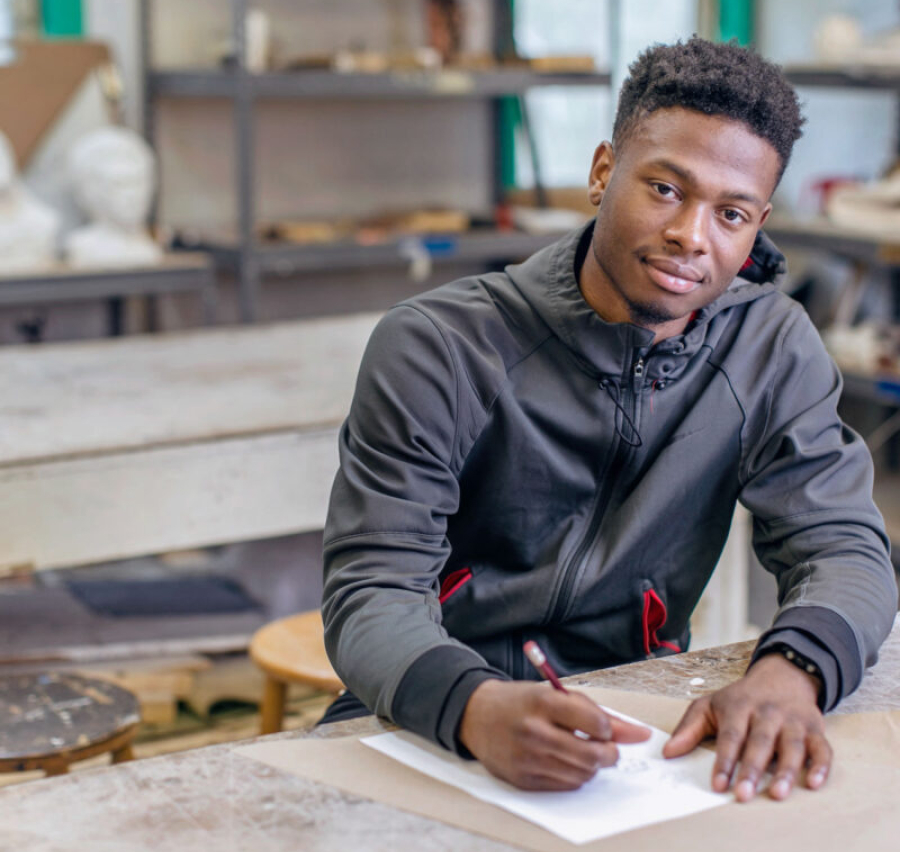 A art student sits in a studio sketching. They seems like they just looked up to get their picture taken.