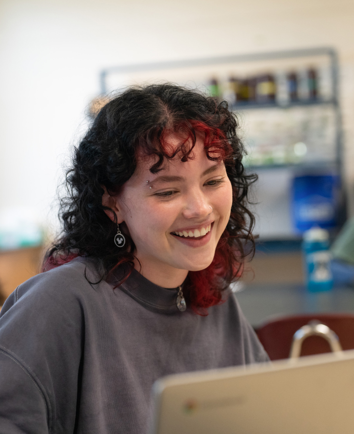 A student sits in a classroom and smiles at their computer.