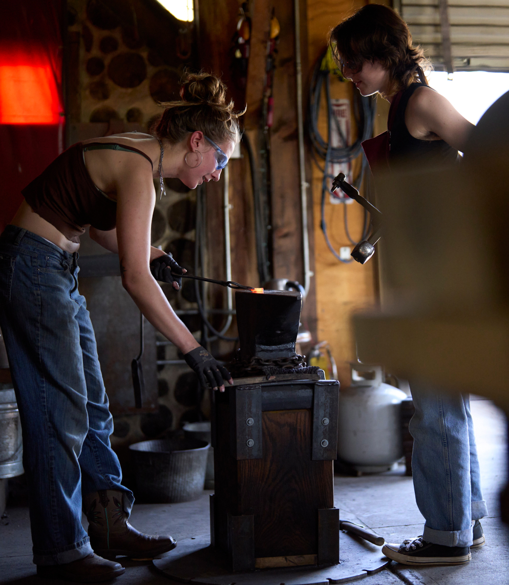 A student uses tongs to hold a molten piece of metal. They are preparing to strike it.