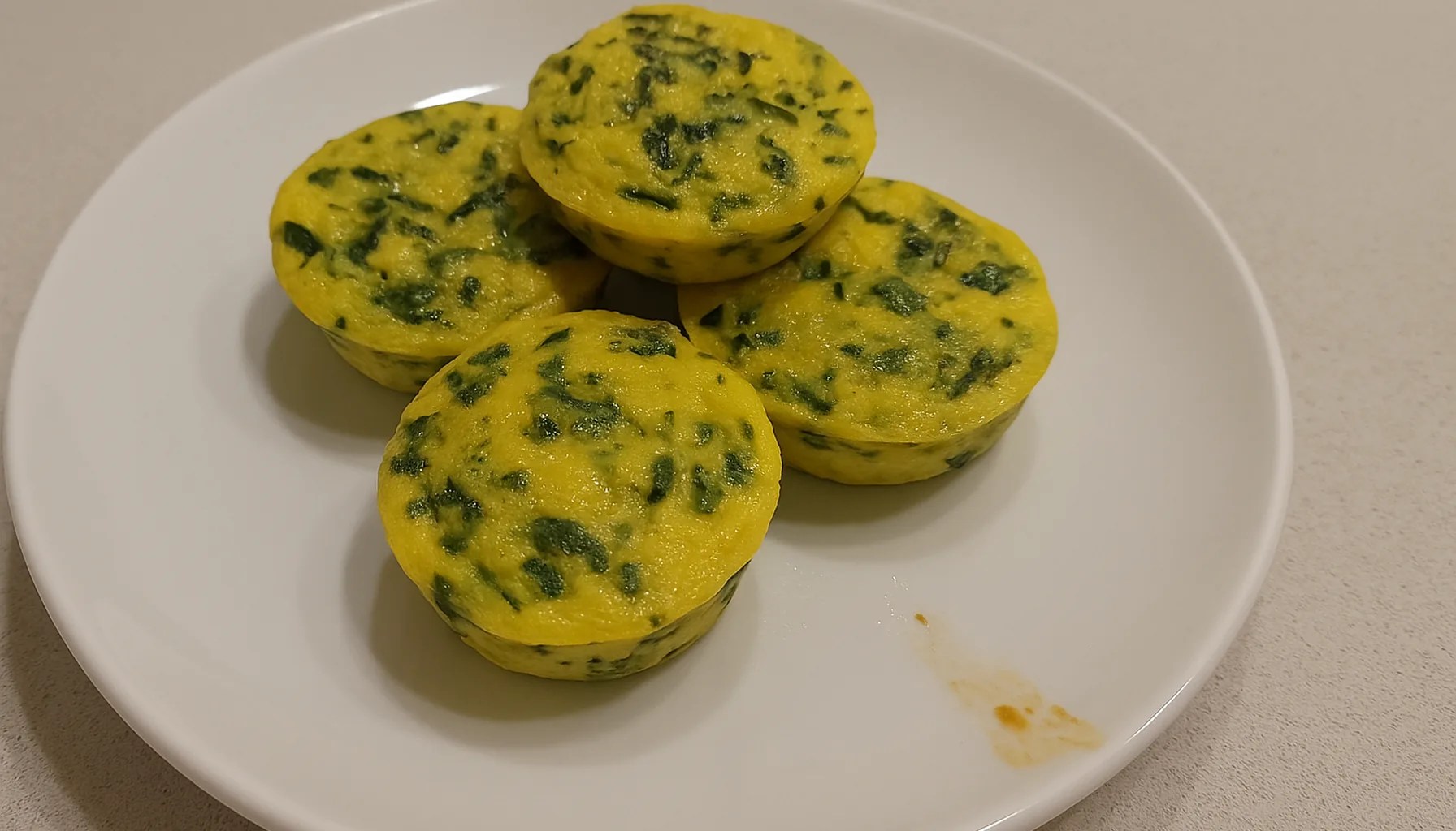 Frittatine soffici allo zafferano e spinaci, con puntini verdi, in un piatto bianco su un piano cucina chiaro — ricetta Zafferù con zafferano in stimmi di Carrù (Piemonte)