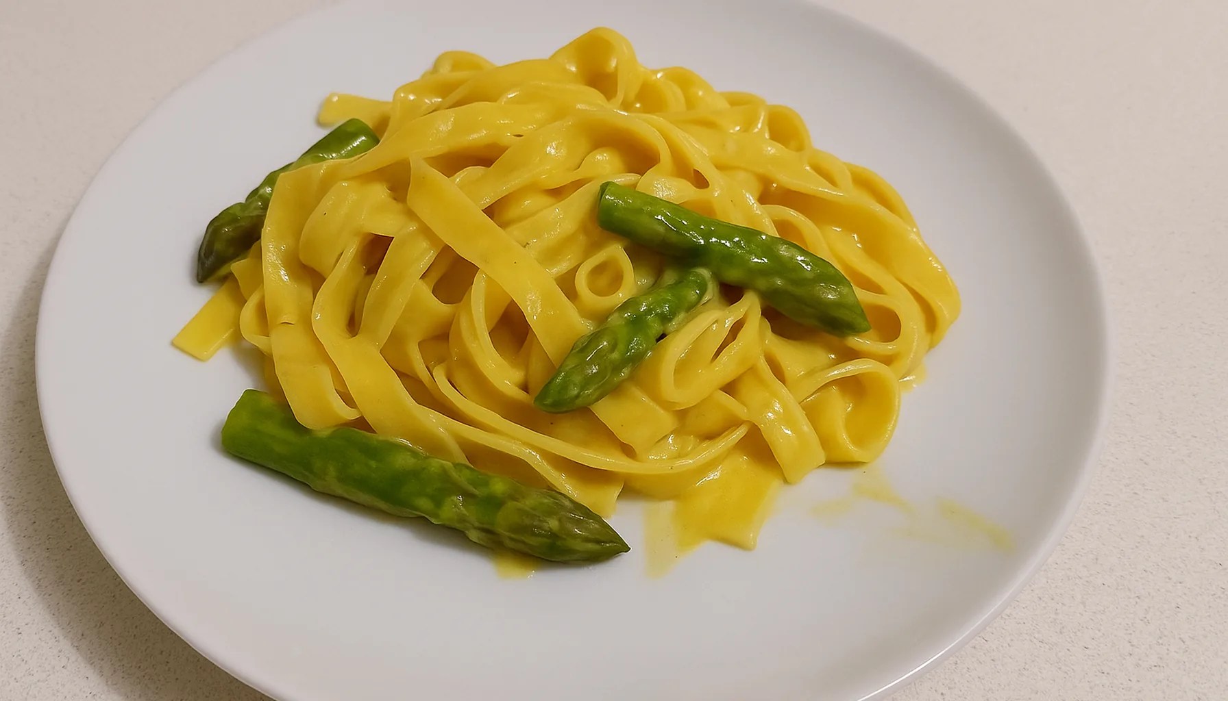 Pasta cremosa allo zafferano con punte di asparagi, in un piatto bianco su un piano cucina chiaro — ricetta Zafferù con zafferano in stimmi di Carrù (Piemonte)