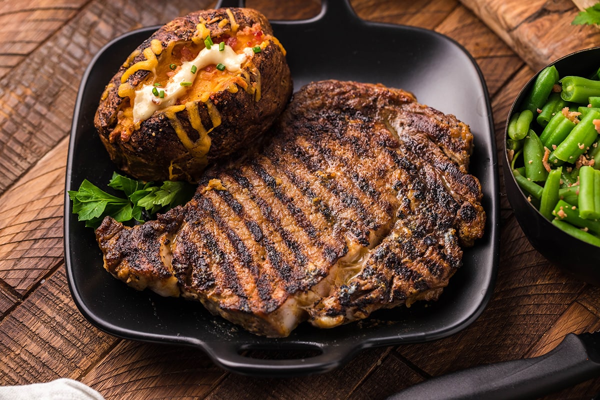 Grilled steak recipes and a loaded baked potato on a black plate, with a side of green beans in a bowl.