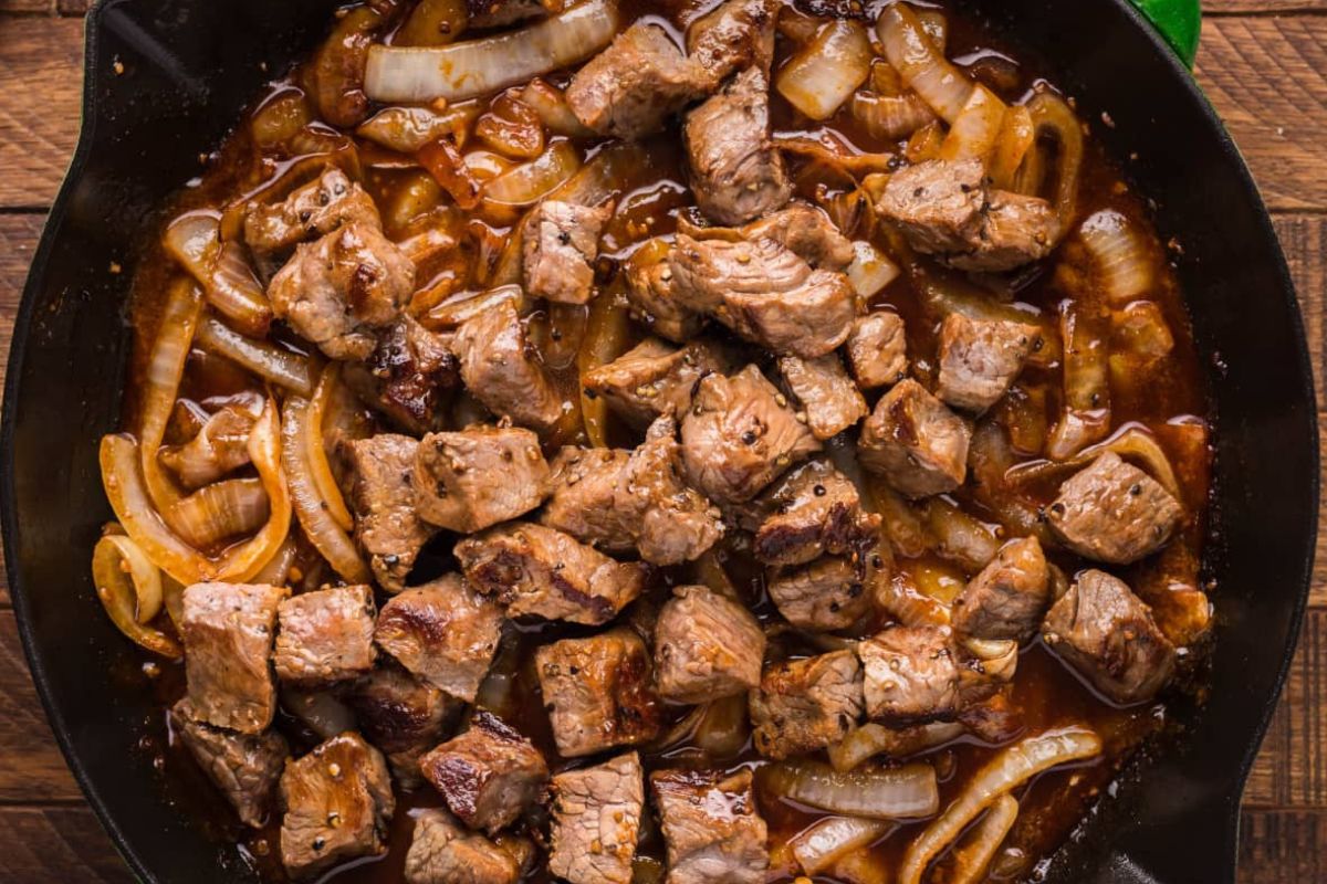 Cast iron skillet with sautéed steak chunks and caramelized onions in a thick sauce.