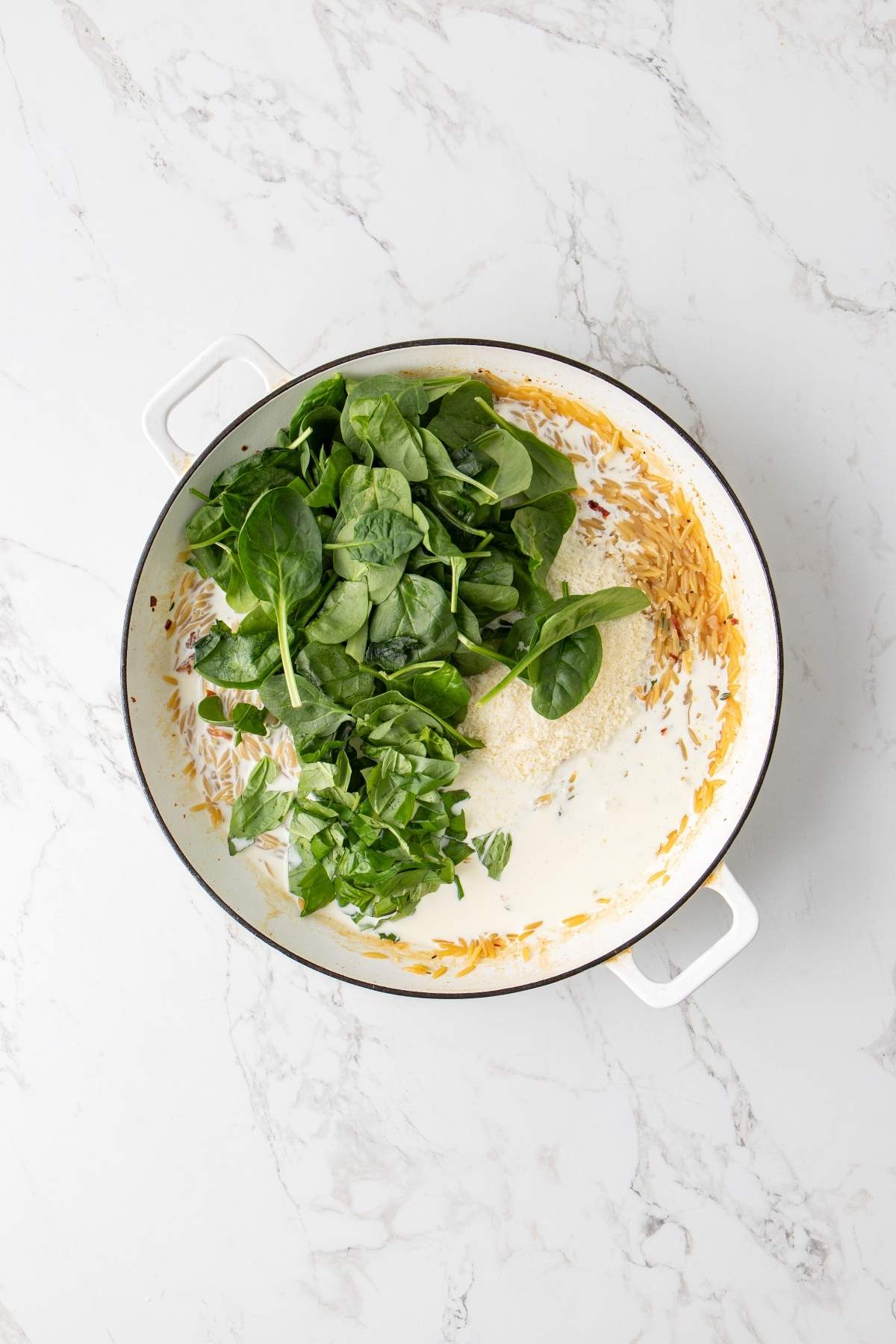 A large white pan with handles contains partially cooked Italian chicken orzo and a pile of fresh spinach, set on a white marble surface.