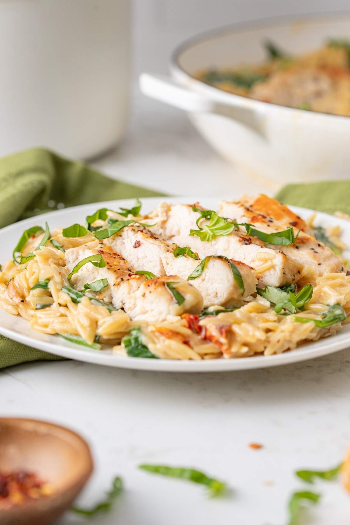 A plate of creamy Tuscan Marry me chicken orzo topped with grilled chicken slices and garnished with chopped green herbs, with a skillet of additional pasta in the background.