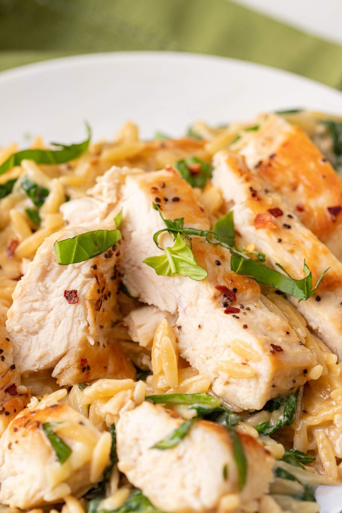 A close-up photo of a creamy Tuscan chicken orzo dish topped with grilled chicken slices and garnished with fresh basil leaves.