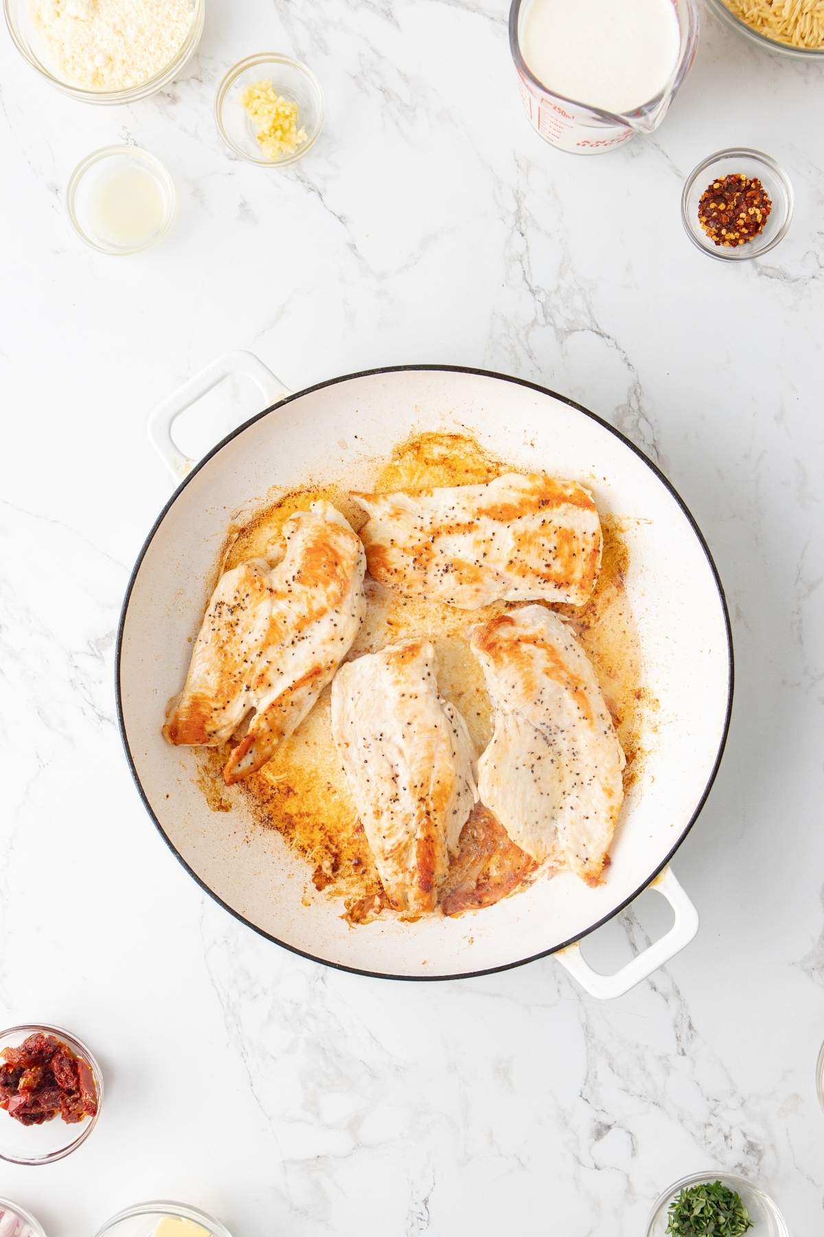 A white skillet contains four seasoned and browned chicken breasts. Surrounding the skillet are various small bowls of ingredients, reminiscent of an Italian chicken orzo recipe, including spices, garlic, and liquids on a marble countertop.