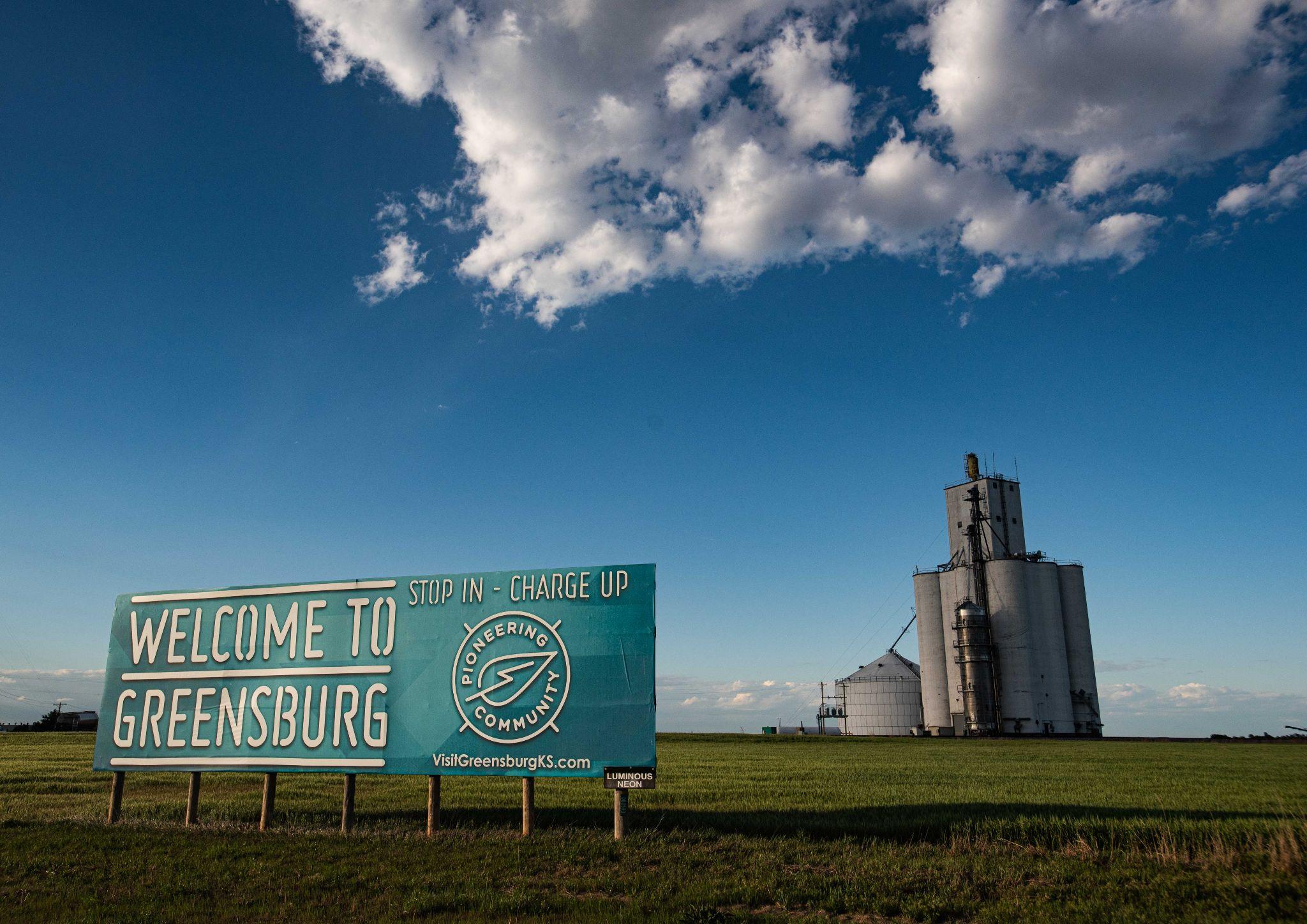 photo of a wide prairie with a grain silo. In the foreground, a billboard reads, "Welcome to Greensburg. Stop in - charge up"