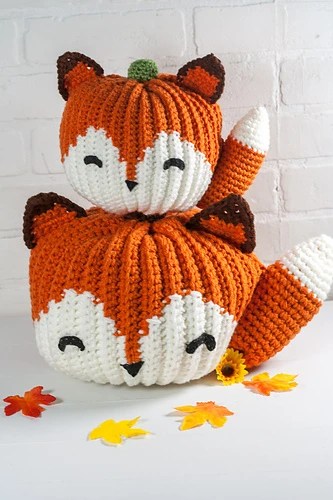 Crochet pumpkin pattern shown in orange and white with the pumpkin made to look like a fox. Includes tail and pointy ears against a white backdrop.