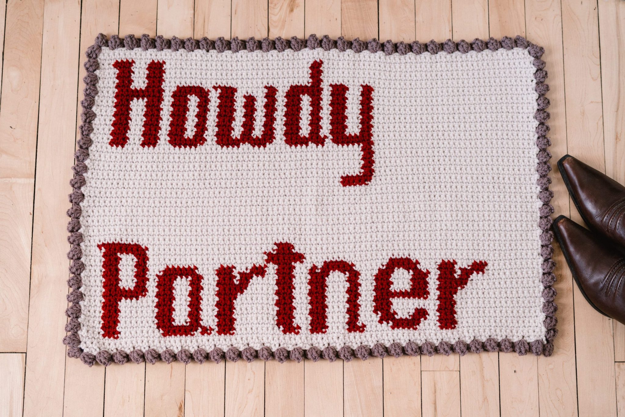 beige crochet rug against hardwood floor. Rug features the text "howdy partner" and has a rope border.