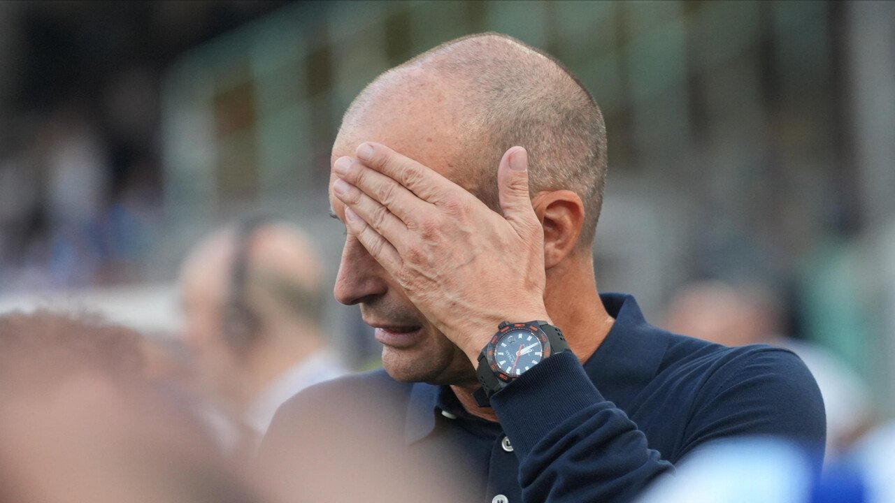 Allenatore in panchina si copre il volto, circondato da persone e spettatori allo stadio.