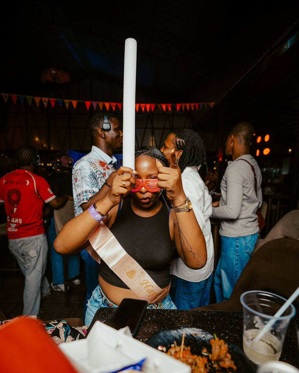 Woman at a lively party wearing a sash, holding a white foam stick and red sunglasses toward the camera.