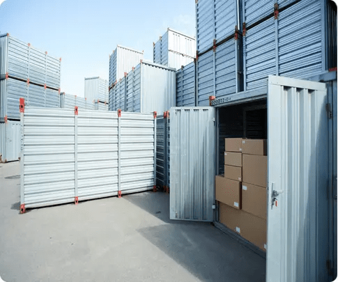 Contenedores metálicos de almacenamiento apilados en un recinto exterior; uno está abierto y contiene varias cajas de cartón.